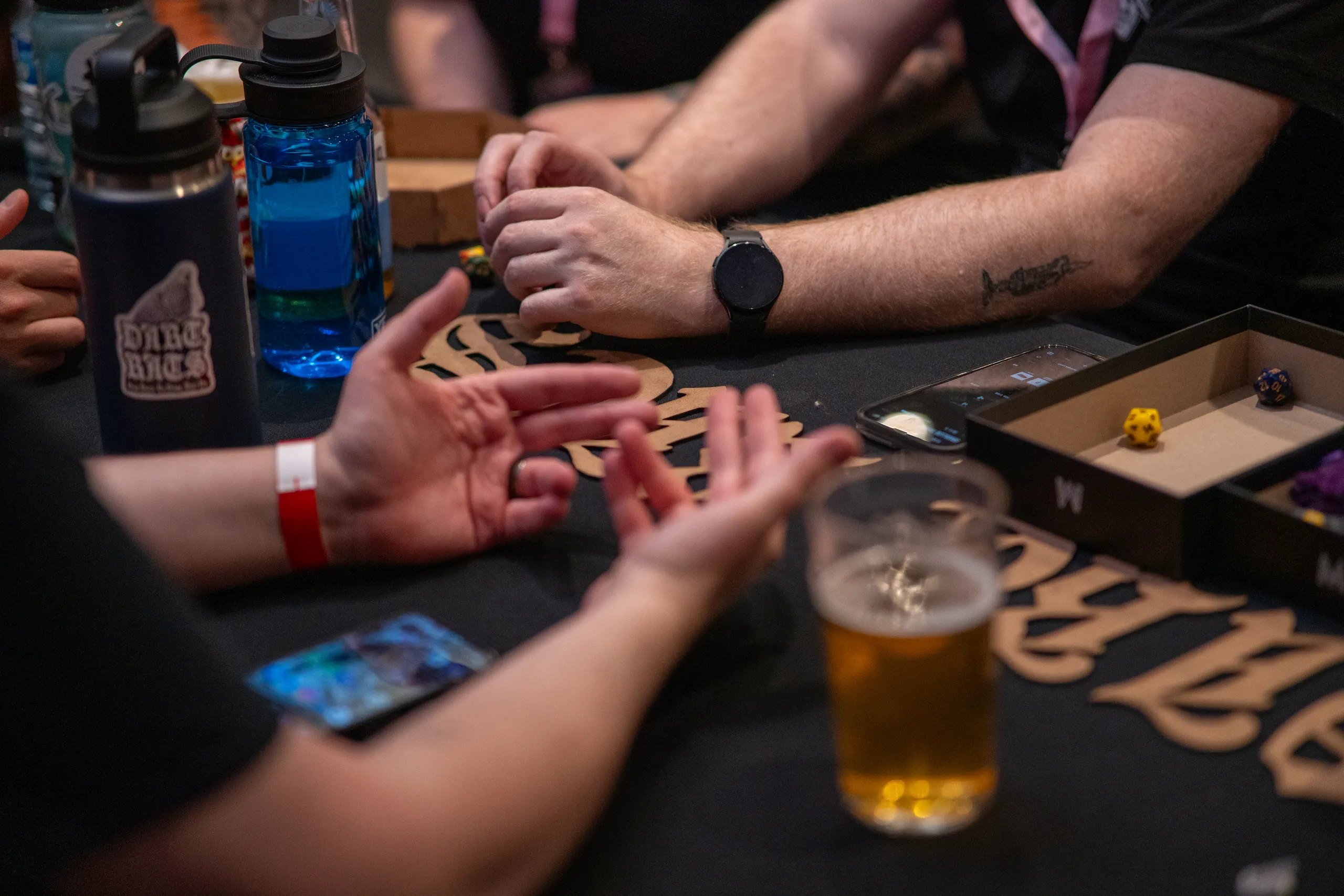 Hands around a table covered in dice, drinks, and wooden game components.